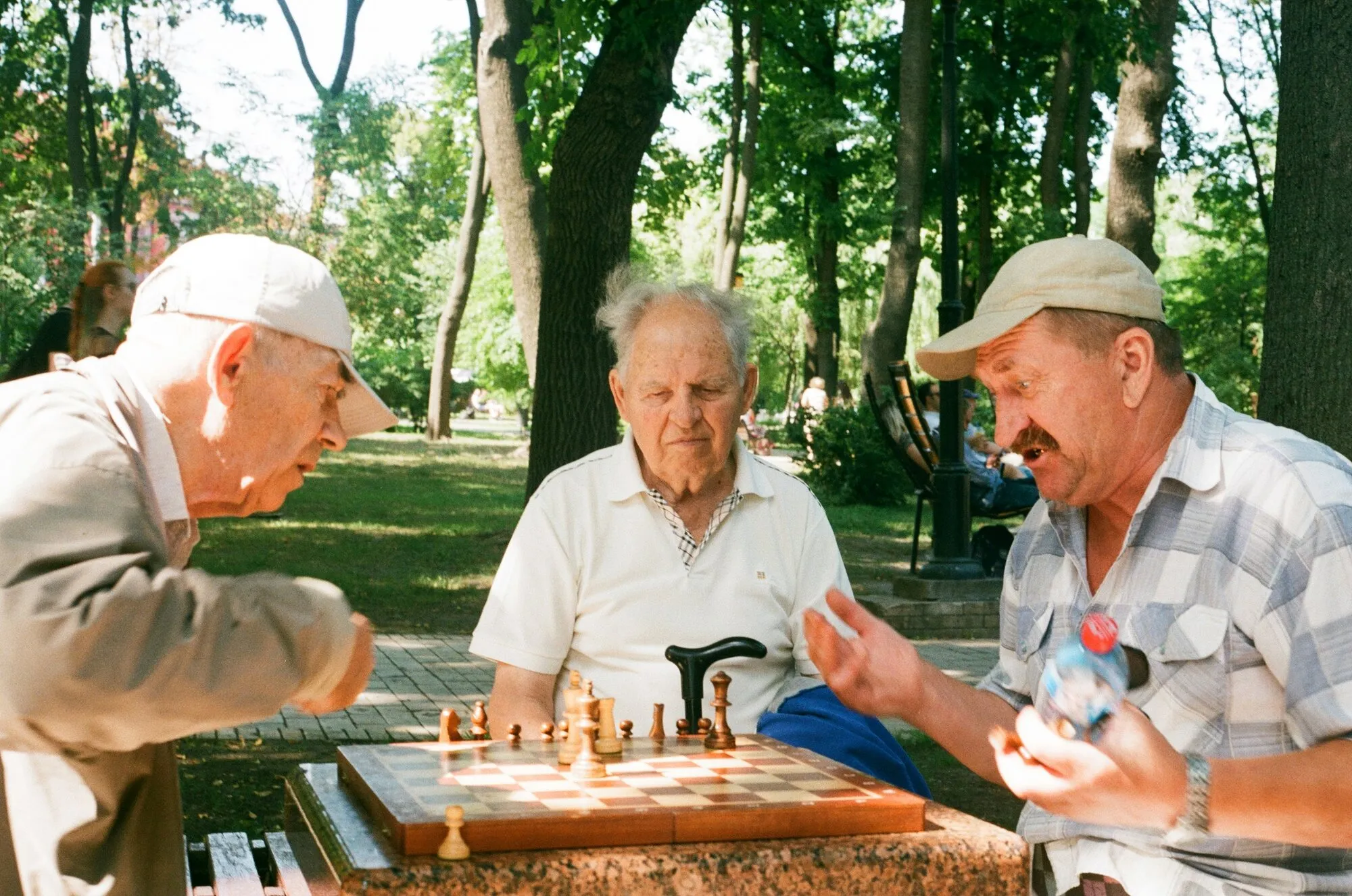Three older men play chess at an outdoor table in a shady park.
