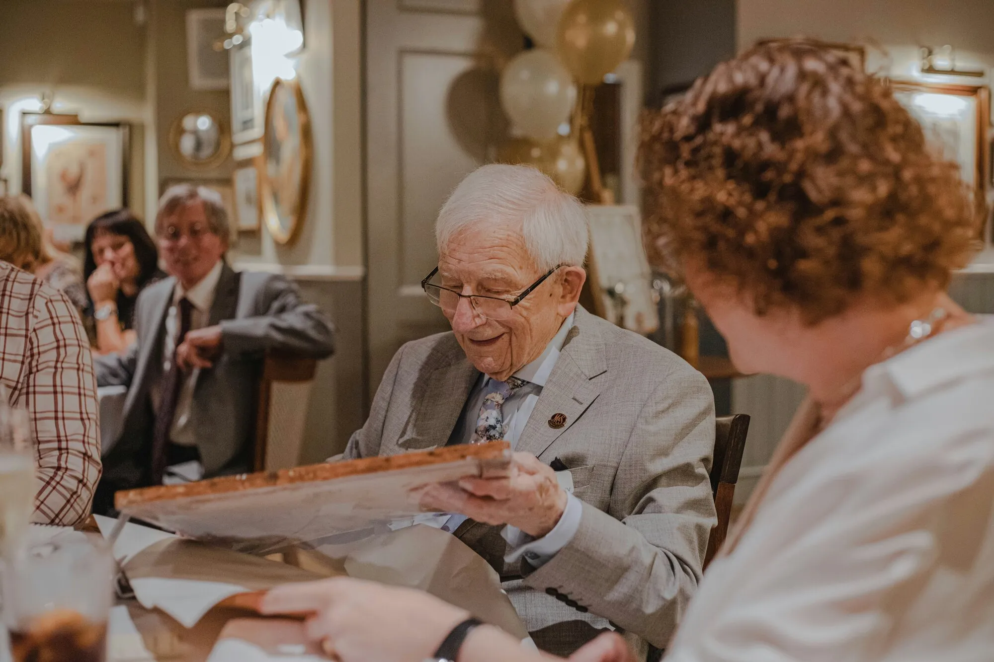 A smiling older man in a suit opens a card at a party with friends.
