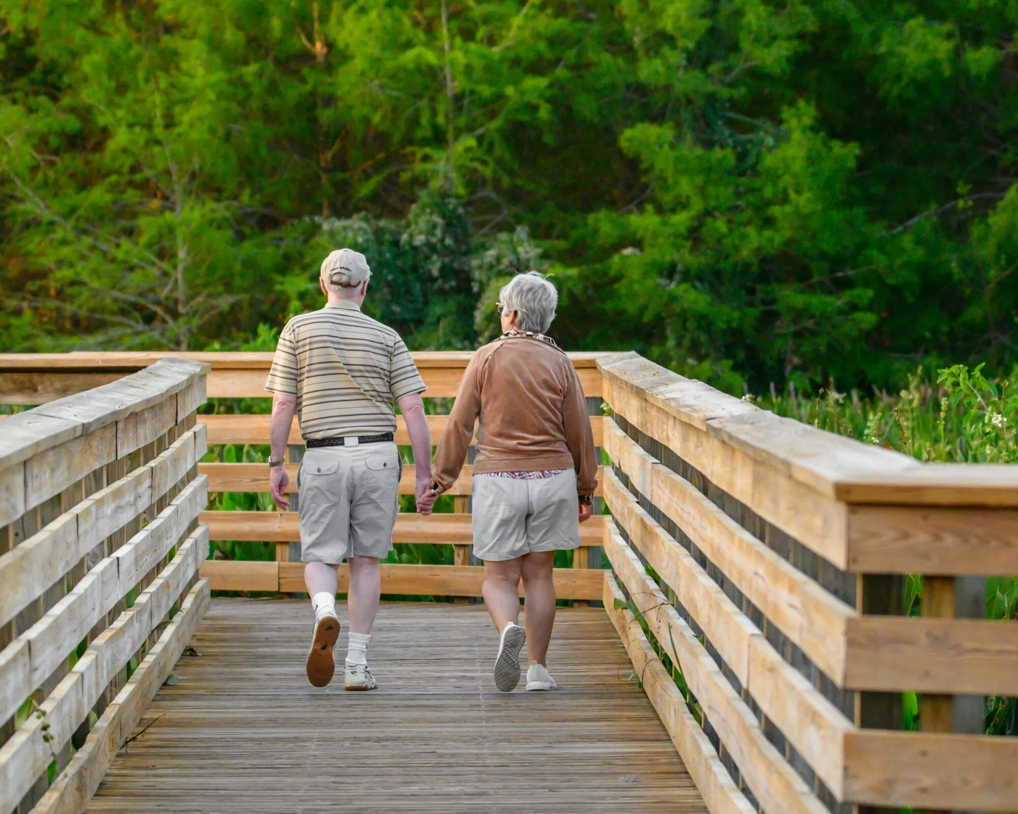 An older couple walks side by side along a wooden boardwalk through green trees.